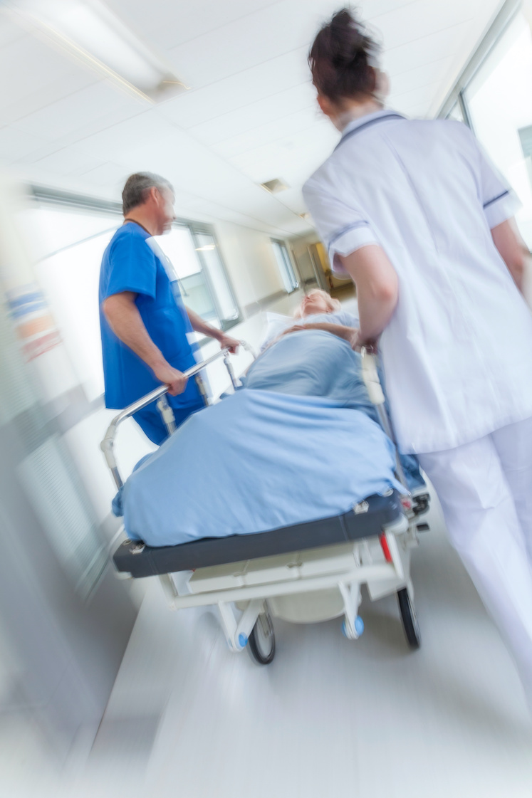A motion blurred photograph of a senior female patient on stretcher or gurney being pushed at speed through a hospital corridor by doctors & nurses to an emergency room