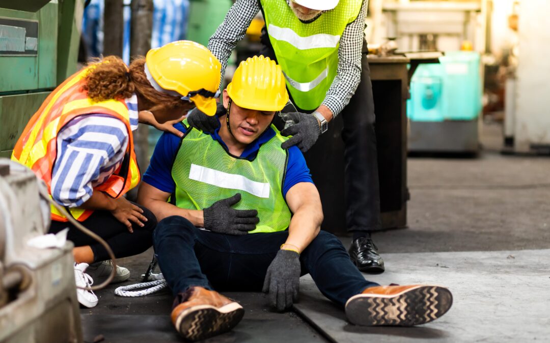 Two workers in hard hats and vests surround a third worker who is expressing that he is hurt.