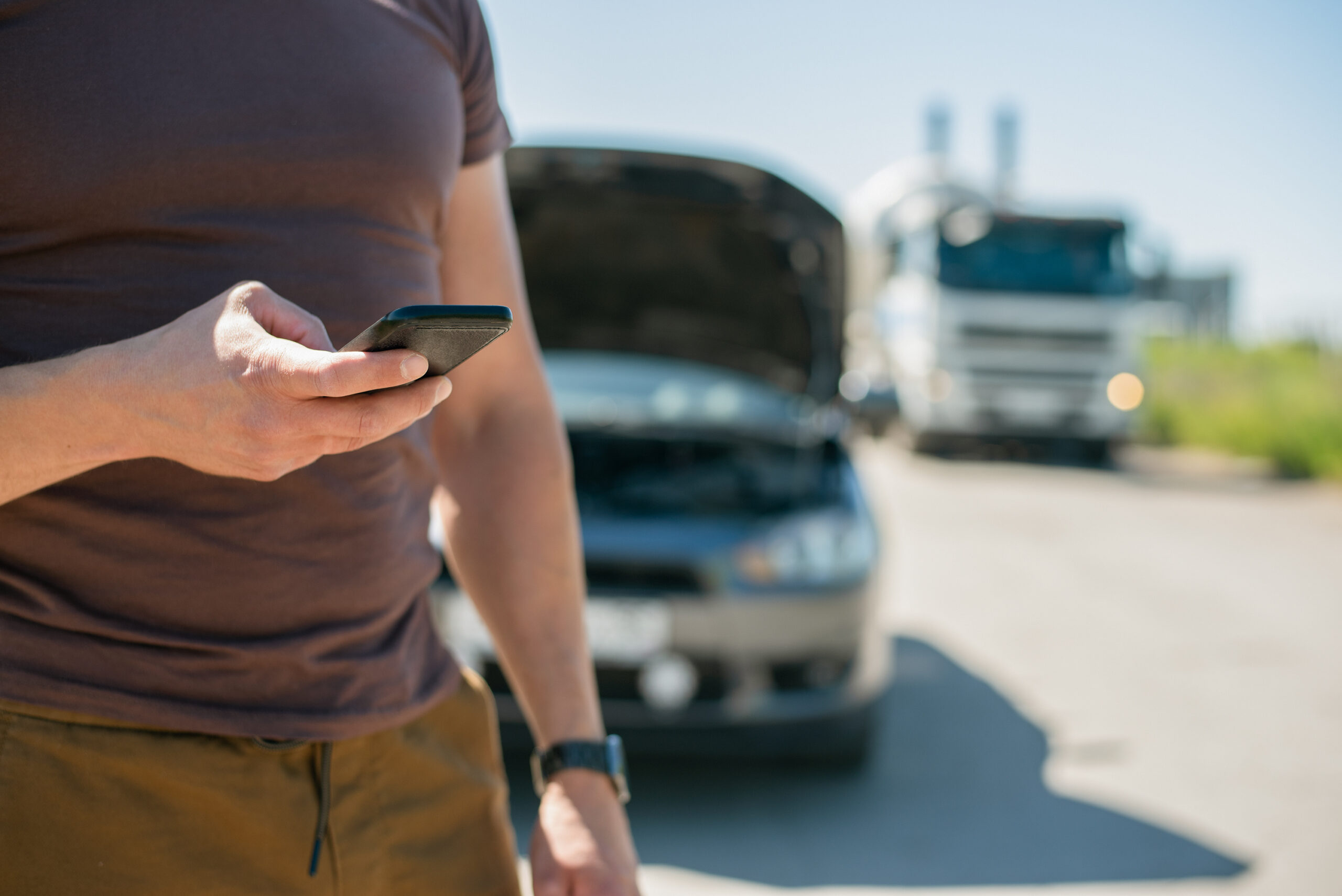 Driver trying to contact someone on his phone while standing outside of his vehicle. Hs vehicle has the hood open and there is an 18-wheeler off to the side of the road behind him.